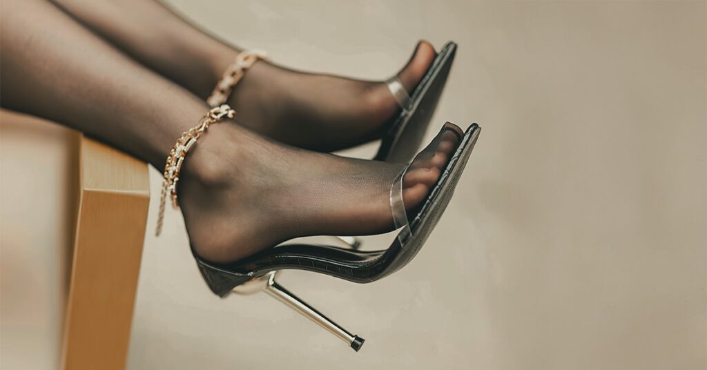 A woman’s foot in black heels and black stockings, laying on a bed to represent the nylon foot fetish