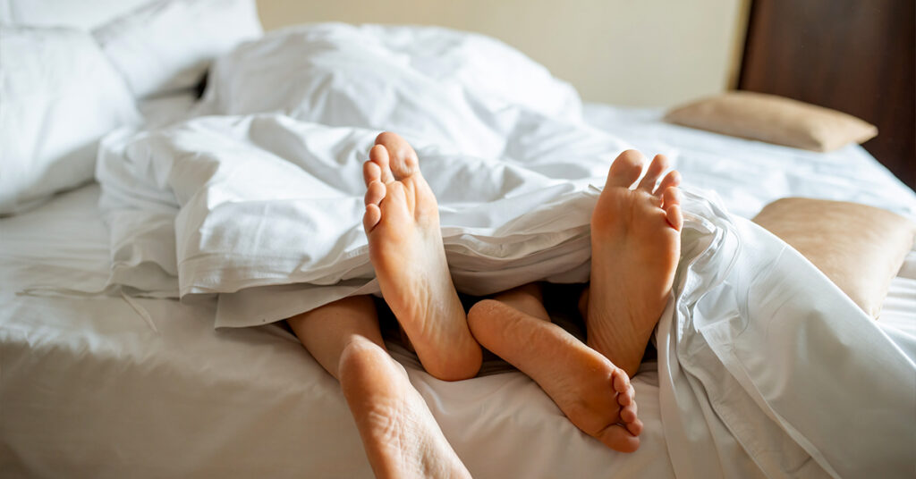 Two pairs of legs entangled in bed, with feet sticking out of the bed to represent sex and climax with feet.